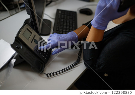 An employee in the office makes a call on a landline phone wearing gloves. Phone close-up. In gray tones. Protection, virus.Stationary phone and headset on desk indoors, closeup. An employee in the office makes a call on a landline phone wearing gloves. Phone close-up. In gray tones. Protection, virus.Stationary phone and headset on desk indoors, closeup. 122298880