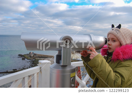 The girl looks through a telescope. A girl looks at ships on the sea horizon with a telescope. The girl looks through a telescope. A girl looks at ships on the sea horizon with a telescope. 122299123