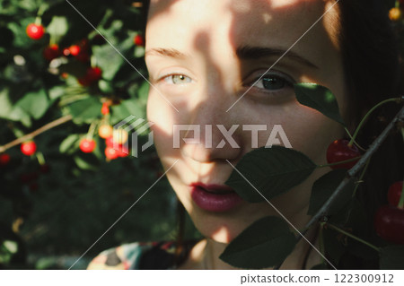 Close up portrait of a young woman plucking ripe red cherries from a tree in an orchard in summer. Harvesting, gardening, farming. Sun rays on a face. 122300912