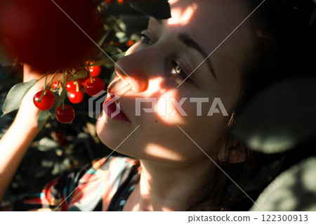 Close up portrait of a young woman plucking ripe red cherries from a tree in an orchard in summer. Harvesting, gardening, farming. Sun rays on a face. 122300913