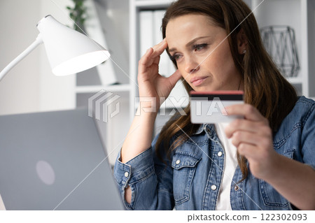 A woman sits at her desk, anxiously shopping online with her credit card at her laptop, feeling overwhelmed and stressed as she contemplates her financial situation and the challenging choices ahead 122302093