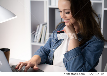 A young woman is happily engaged in the activity of online shopping from the comfort of her own home, enjoying the convenience it brings while using her laptop at a cozy desk in her trendy workspace 122302104