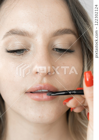 A beautiful young woman transforms under the spell of makeup created by a makeup artist. The face reflects the uniqueness and natural appeal, emphasized by the graceful touches of cosmetics. 122302124