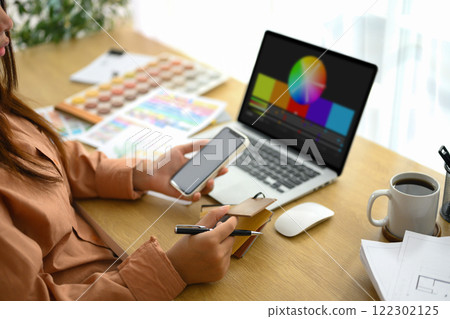 Female designer holding smartphone working with material and color samples at workplace Female designer holding smartphone working with material and color samples at workplace 122302125