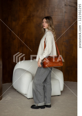 beautiful girl in gray pants and beige sweater posing with a small shiny brown leather bag 122302295