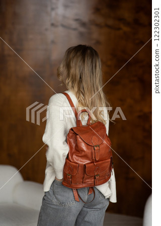 beautiful girl in gray pants and beige sweater posing with a small shiny brown leather backpack 122302301