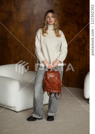beautiful girl in gray pants and beige sweater posing with a small shiny brown leather backpack 122302302