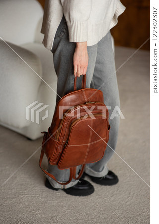 beautiful girl in gray pants and beige sweater posing with a small shiny brown leather backpack beautiful girl in gray pants and beige sweater posing with a small shiny brown leather backpack 122302307