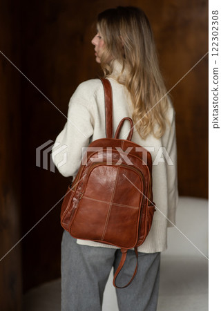 beautiful girl in gray pants and beige sweater posing with a small shiny brown leather backpack beautiful girl in gray pants and beige sweater posing with a small shiny brown leather backpack 122302308