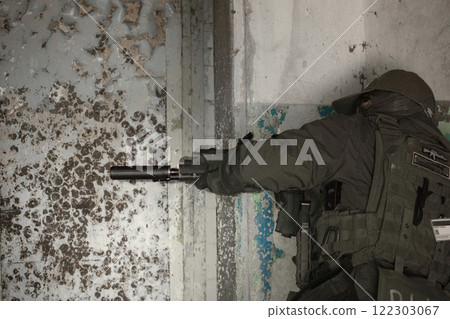 A tactical soldier in full gear takes cover behind a wall in an abandoned building, aiming a silenced rifle. A tactical soldier in full gear takes cover behind a wall in an abandoned building, aiming a silenced rifle. 122303067