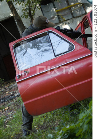 A tactical scene featuring a person in military gear aiming a rifle from behind cover provided by an old car. A tactical scene featuring a person in military gear aiming a rifle from behind cover provided by an old car. 122303074