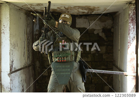 A tactical operator dressed in military gear and a cap stands inside an abandoned building, holding a rifle. 122303089