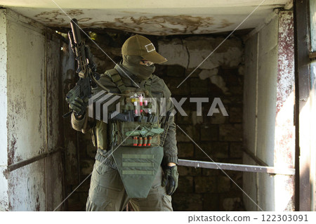 A tactical operator dressed in military gear and a cap stands inside an abandoned building, holding a rifle. A tactical operator dressed in military gear and a cap stands inside an abandoned building, holding a rifle. 122303091