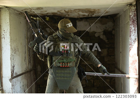 A tactical operator dressed in military gear and a cap stands inside an abandoned building, holding a rifle. 122303093