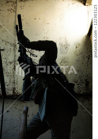 A tactical operator in military gear ascends a staircase in a dimly lit, abandoned building, aiming a rifle upwards. A tactical operator in military gear ascends a staircase in a dimly lit, abandoned building, aiming a rifle upwards. 122303125