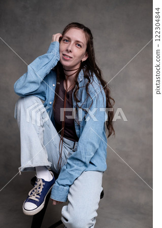 beautiful and long-haired woman in stylish clothes posing in a photo studio and smiling. a model in braces sits on a chair and poses in denim and sneakers on a gray photo background. 122304844
