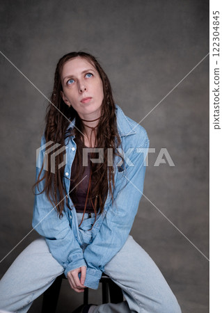 a beautiful and long-haired woman with big blue eyes in stylish clothes posing in a photo studio. the model sits on a chair and poses in denim clothes on a gray photo background. 122304845