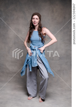 a beautiful and long-haired woman in a hat and stylish clothes poses in a photo studio and smiles. model stands and poses in sportswear and barefoot on a gray photo background a beautiful and long-haired woman in a hat and stylish clothes poses in a photo studio and smiles. model stands and poses in sportswear and barefoot on a gray photo background 122304847