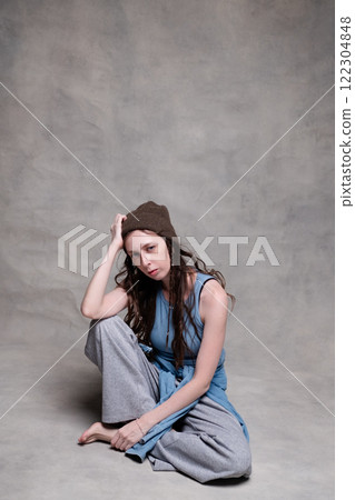 beautiful and long-haired woman in a hat and stylish clothes poses in a photo studio with a serious face. model sitting and posing in sportswear and barefoot on a gray photo background beautiful and long-haired woman in a hat and stylish clothes poses in a photo studio with a serious face. model sitting and posing in sportswear and barefoot on a gray photo background 122304848