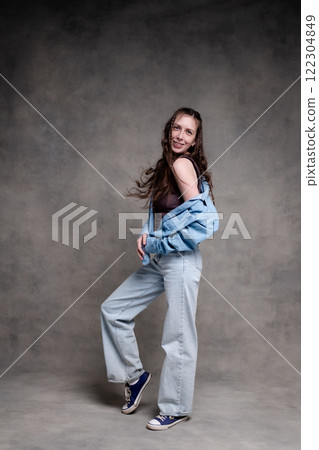 beautiful and long haired woman in stylish clothes posing in a photo studio and smiling. the model stands and poses in denim and sneakers on a gray photo background. 122304849