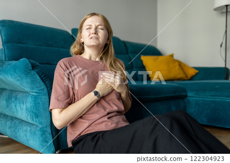 Woman sitting with strong chest pain and hand touching her chest while having trouble at home. Heart attack concept or heart failure symptom. Pressure in the chest. Woman sitting with strong chest pain and hand touching her chest while having trouble at home. Heart attack concept or heart failure symptom. Pressure in the chest. 122304923
