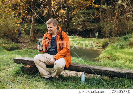 Stylish man with backpack holding smart phone and relaxing in nature. Full body photo. Hiking active male lifestyle siting on tree trunk outdoors. High quality photo. Adventures. 122305278
