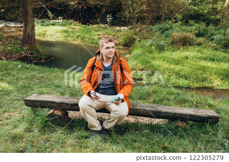 Stylish man with backpack holding smart phone and relaxing in nature. Full body photo. Hiking active male lifestyle siting on tree trunk outdoors. High quality photo. Adventures. 122305279
