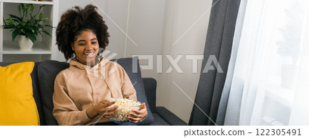 Banner African American woman holds a bowl of popcorn while enjoying a television show. Bright smile radiates happiness and relaxation in a cozy home. TV, series and streaming service concept. Copy Banner African American woman holds a bowl of popcorn while enjoying a television show. Bright smile radiates happiness and relaxation in a cozy home. TV, series and streaming service concept. Copy 122305491