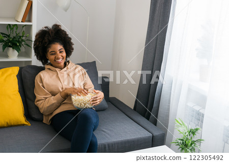 Young African American woman holds a bowl of popcorn while enjoying a television show. Bright smile radiates happiness and relaxation in a cozy home. TV, series and streaming service concept. Copy Young African American woman holds a bowl of popcorn while enjoying a television show. Bright smile radiates happiness and relaxation in a cozy home. TV, series and streaming service concept. Copy 122305492