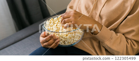 Banner close up of african american female hand taking popcorn from big glass bowl during watching movie at home. Copy space 122305499