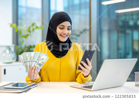 Joyful and successful woman in hijab sitting at desk inside office. Arab businesswoman holding money cash dollars and phone in hands, received online loan using banking app. Joyful and successful woman in hijab sitting at desk inside office. Arab businesswoman holding money cash dollars and phone in hands, received online loan using banking app. 122305833