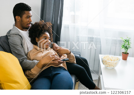 African American couple relaxes on a cozy sofa, sharing a joyful moment while watching a show with a bowl of popcorn. Essence of quality time and comfort in a stylish modern living room 122306157