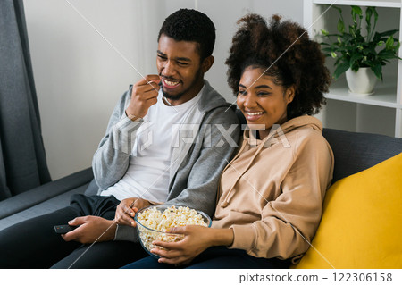 African American couple wife husband watching movie. Family pastime and leisure, watch television, rest. Relaxed man and woman watching TV series together sitting on couch at home enjoying eating 122306158