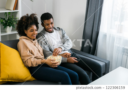 African American couple wife husband watching movie. Family pastime and leisure, watch television, rest. Relaxed man and woman watching TV series together sitting on couch at home enjoying eating 122306159