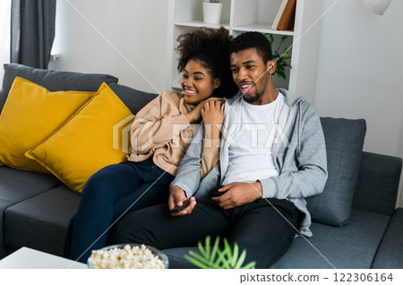 African American couple wife husband watching movie. Family pastime and leisure, watch television, rest. Relaxed man and woman watching TV series together sitting on couch at home enjoying eating African American couple wife husband watching movie. Family pastime and leisure, watch television, rest. Relaxed man and woman watching TV series together sitting on couch at home enjoying eating 122306164