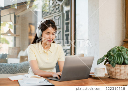 A young woman wearing headphones works on a laptop in a bright, cozy home office with natural light and indoor plants. 122310316