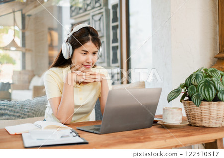 A young woman wearing headphones, focused on her laptop in a cozy home office setting with natural light and greenery. 122310317