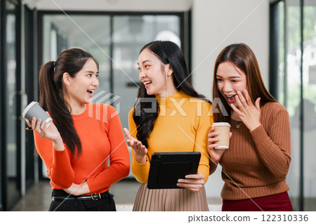Three women in a bright office, chatting and holding coffee cups, dressed in casual attire, showcasing friendship and modern work culture. 122310336