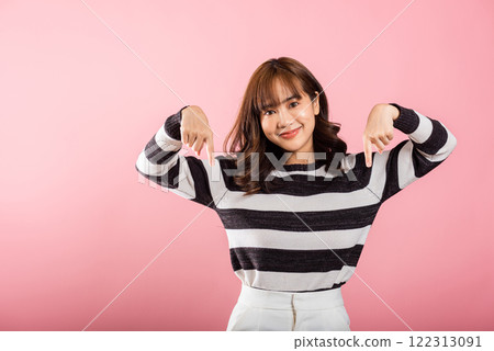 An Asian lady stands in a studio portrait, smiling and making a two-finger pointing gesture downwards. Isolated on pink, ideal for advertisements and promotions. An Asian lady stands in a studio portrait, smiling and making a two-finger pointing gesture downwards. Isolated on pink, ideal for advertisements and promotions. 122313091