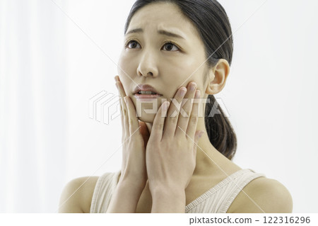 A young woman with a troubled face holding both cheeks on a white background, skin irritation 122316296