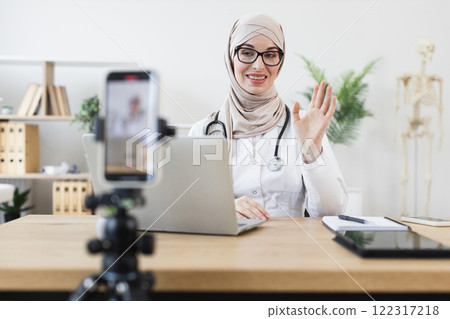 Muslim woman doctor recording video lesson on mobile phone. Young female in hijab shares medical advice, sitting at desk in modern office. Muslim woman doctor recording video lesson on mobile phone. Young female in hijab shares medical advice, sitting at desk in modern office. 122317218