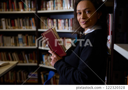 A woman in a library, holding a red book and gazing thoughtfully at the camera. She is surrounded by shelves filled with books, reflecting a moment of study or contemplation. A woman in a library, holding a red book and gazing thoughtfully at the camera. She is surrounded by shelves filled with books, reflecting a moment of study or contemplation. 122320328