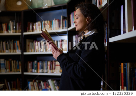 A contemplative woman standing by bookshelves in a library, reading a book. She is engrossed in her reading, creating a peaceful scene that reflects knowledge, learning, and personal reflection. A contemplative woman standing by bookshelves in a library, reading a book. She is engrossed in her reading, creating a peaceful scene that reflects knowledge, learning, and personal reflection. 122320329