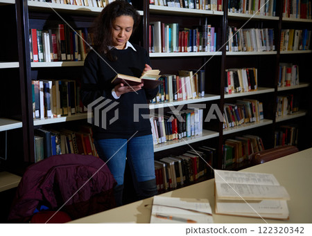 A woman stands in a library, engrossed in a book, surrounded by shelves of literature, and studying at a desk. A woman stands in a library, engrossed in a book, surrounded by shelves of literature, and studying at a desk. 122320342