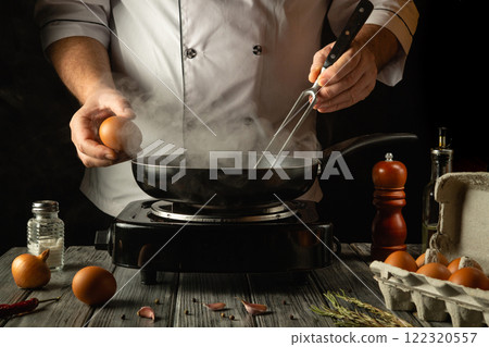 A chef prepares to create a tasty dish by cracking fresh eggs into a hot skillet. Steam rises as culinary magic begins at an early hour, accompanied by rustic ingredients on the table A chef prepares to create a tasty dish by cracking fresh eggs into a hot skillet. Steam rises as culinary magic begins at an early hour, accompanied by rustic ingredients on the table 122320557