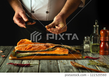 A skilled chef seasons a fresh salmon fillet with vibrant spices, blending rich colors and aromas in a rustic kitchen. The warm atmosphere highlights culinary creativity and tradition 122320560