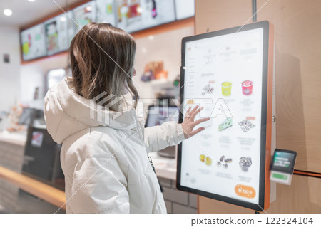 A 30 -35-year-old woman in a white winter jacket orders food through the screen menu at a fast food restaurant. A customer is utilizing a digital kiosk designed specifically for the purpose of A 30 -35-year-old woman in a white winter jacket orders food through the screen menu at a fast food restaurant. A customer is utilizing a digital kiosk designed specifically for the purpose of 122324104