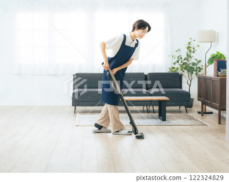 A middle-aged woman wearing an apron vacuuming the floor of a room 122324829