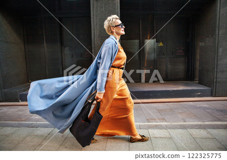 Stylish Woman Walking City Street In Fashionable Attire, Wearing Long Beige Silk Dress, Blue Cape Coat, Carrying Large Black Handbag, Sunglasses And Trendy Loafers Stylish Woman Walking City Street In Fashionable Attire, Wearing Long Beige Silk Dress, Blue Cape Coat, Carrying Large Black Handbag, Sunglasses And Trendy Loafers 122325775