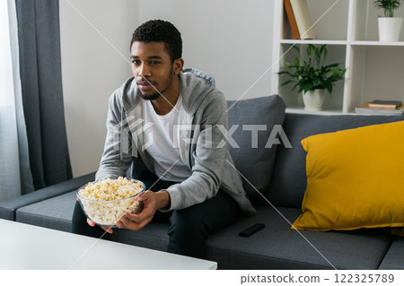 African American man sits alone on the couch and enjoying popcorn while watching television and streaming service. Copy space African American man sits alone on the couch and enjoying popcorn while watching television and streaming service. Copy space 122325789